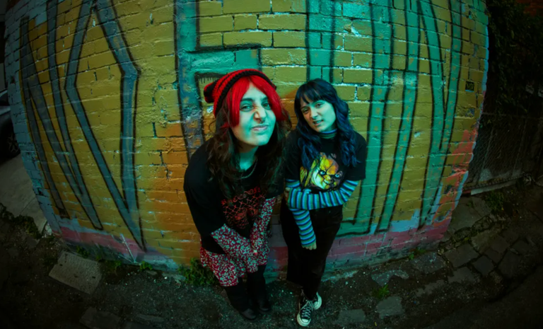 Indie rock duo Teenage Joans posing outdoors in front of a colourful graffiti brick wall in a wide-angle promotional photo.